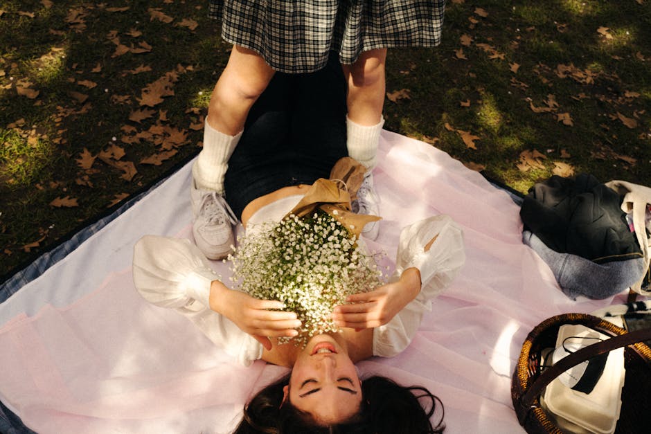 A young woman with long dark hair and fair skin lies on a pink tulle blanket outdoors on a grassy area scattered with brown fallen leaves. She is smiling, holding a bouquet of baby's breath flowers wr