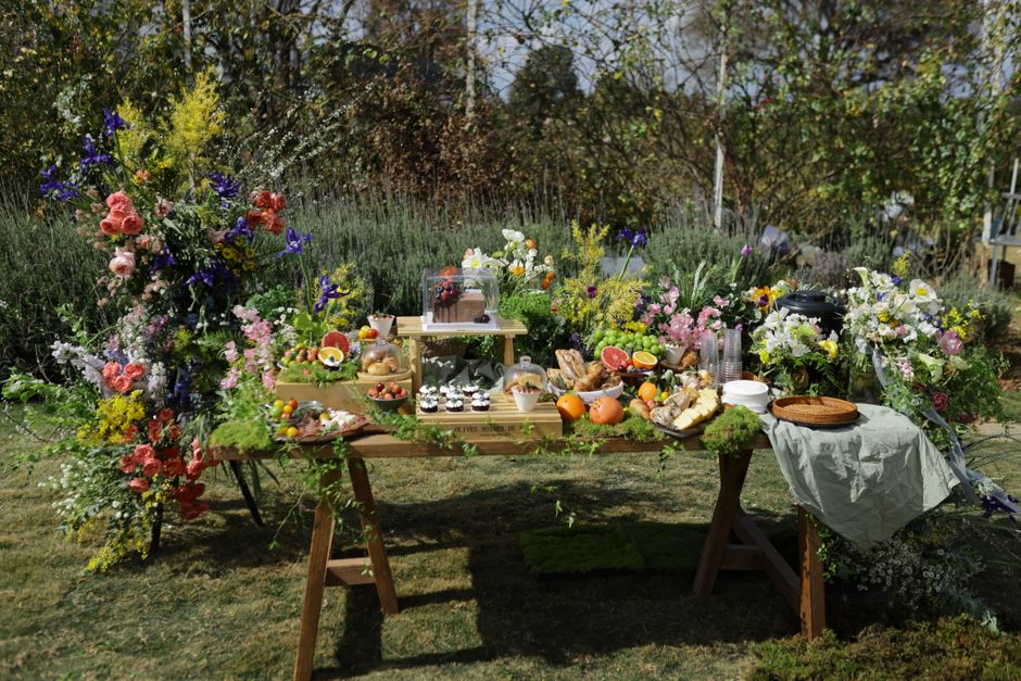 A vibrant outdoor flower display featuring a variety of fresh floral arrangements and bouquets, predominantly in shades of pink, purple, white, and yellow, with some greenery accents. The flowers incl