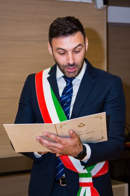 A man dressed in a dark blue suit with a blue and white striped tie, wearing a tricolored sash in red, white, and green, stands indoors against a wooden-paneled wall. He is holding an open envelope or